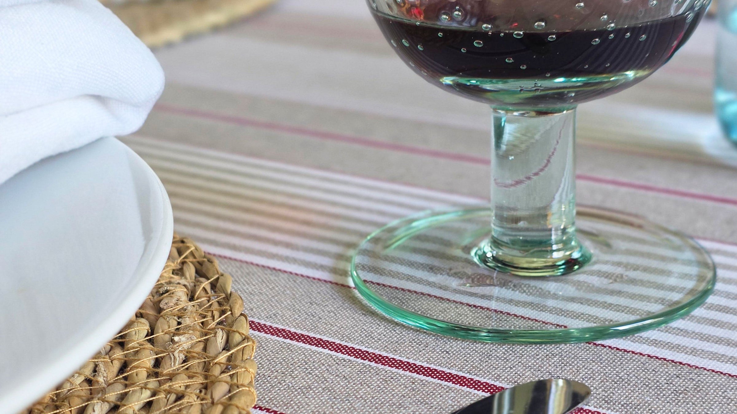 Green recycled glass wine glass containing red wine on a red striped tablecloth next to a placemat with white plate and white napkin