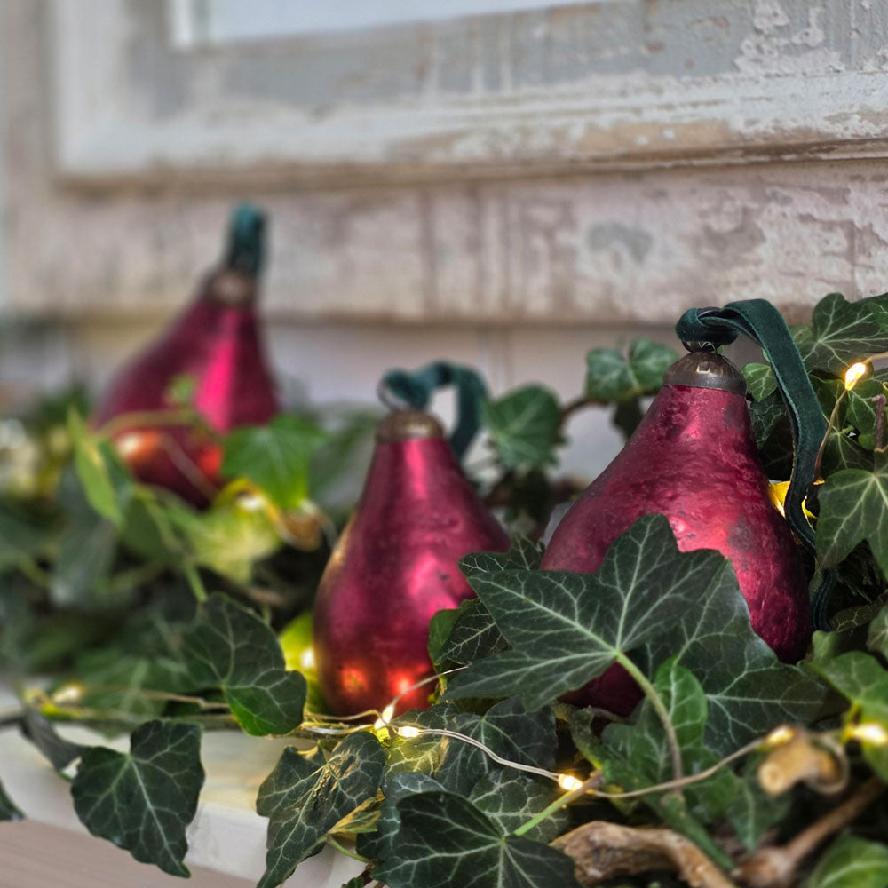 Three metallic burgundy pear-shaped Christmas ornaments with dark green ribbons and warm white fairy lights, nestled among green ivy leaves on a rustic white windowsill.