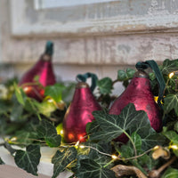 Three metallic burgundy pear-shaped Christmas ornaments with dark green ribbons and warm white fairy lights, nestled among green ivy leaves on a rustic white windowsill.