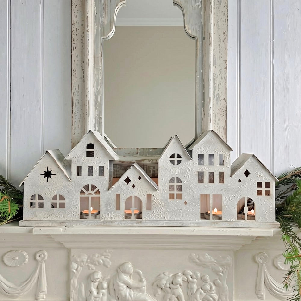 A decorative white house-shaped ornament with six small cottage roofs, lit with tea lights inside, is displayed on a white carved mantelpiece in front of a mirror.