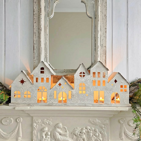 A white distressed house decoration with glowing tea light windows, displayed on a white carved mantelpiece in front of a mirror.