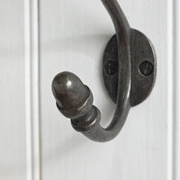 A close-up of a dark metal hook with a decorative ribbed tip, mounted on a white paneled surface.
