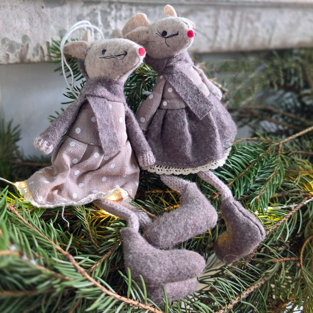 Two handmade felt mouse ornaments dressed in muted grayish-purple and taupe dresses with white polka dots, posed among green pine branches with warm string lights.