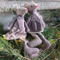 Two handmade felt mouse ornaments dressed in muted grayish-purple and taupe dresses with white polka dots, posed among green pine branches with warm string lights.