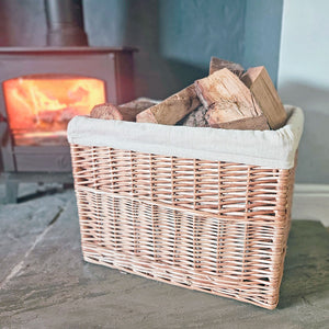 Small wicker log basket with linen lining filled with logs next to lit log burner.