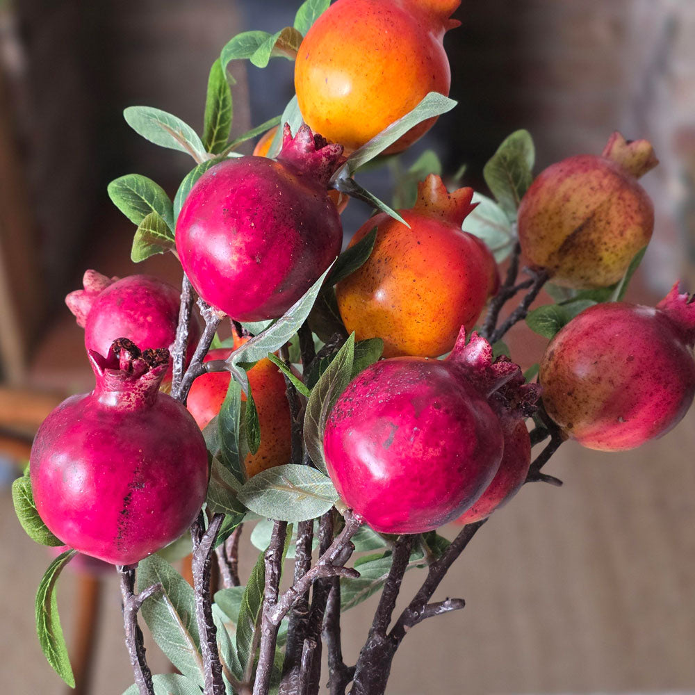 Artificial pomegranate branch arrangement with glossy red-pink fruits, orange-red fruits, and green leaves on dark branches, photographed close-up against a softly blurred neutral background.