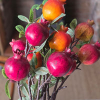 Artificial pomegranate branch arrangement with glossy red-pink fruits, orange-red fruits, and green leaves on dark branches, photographed close-up against a softly blurred neutral background.