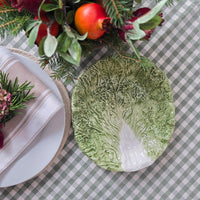 A ceramic plate shaped like a cabbage leaf, featuring realistic textured green and white leaf details, styled on a green and white checkered tablecloth with holiday decorations.