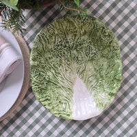 A round ceramic plate sculpted to look like a cabbage leaf, featuring realistic textured green leaves and a white core, displayed on a green and white checkered tablecloth.