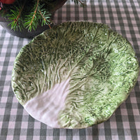 A ceramic plate shaped like a cabbage leaf, featuring textured green outer leaves and a pale white inner core, set on a black and white checkered tablecloth.
