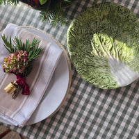 A styled tabletop scene with a round green leaf-shaped plate on a green-and-white checkered tablecloth, alongside a folded cream napkin, pine sprigs, and a small floral boutonniere.