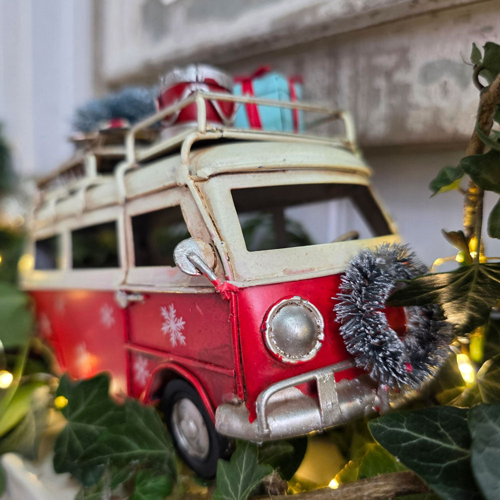 A vintage-style red and cream toy camper van decorated for Christmas, with snowflake decals, a silver bumper, a tinsel wreath on the front, and gift boxes on the roof, surrounded by green ivy and warm fairy lights.
