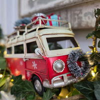A vintage-style red and cream toy camper van decorated for Christmas, with snowflake decals, a silver bumper, a tinsel wreath on the front, and gift boxes on the roof, surrounded by green ivy and warm fairy lights.