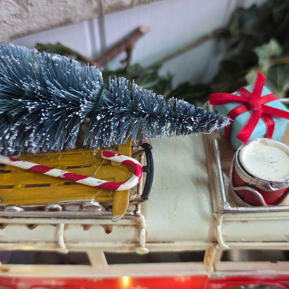 A close-up of a miniature Christmas scene featuring a small blue frosted tree on a yellow sleigh, a red-and-white striped candy cane, a light blue gift box with a red ribbon, and a small tin canister on a cream-colored train car.