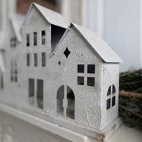 A small white textured house-shaped decorative object with cutout windows and doors, photographed close up on a white surface with a blurred background and greenery at the side.