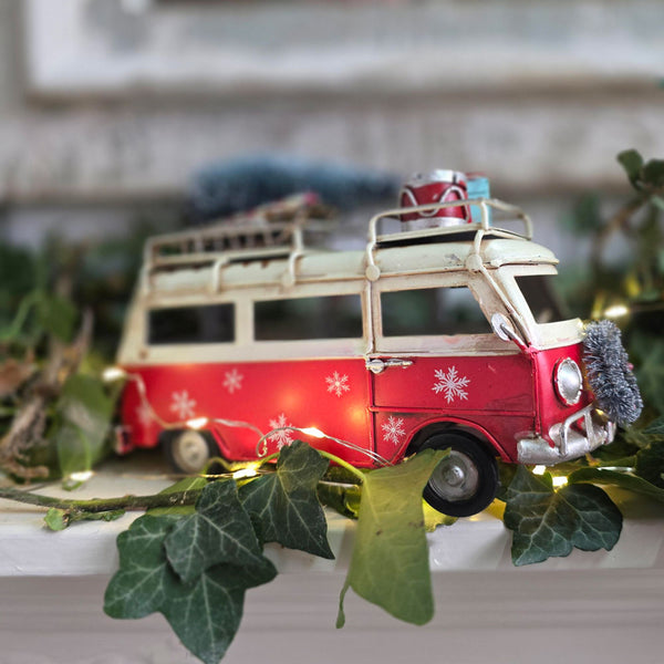 A vintage-style toy camper van decoration in red and cream, decorated with white snowflakes, a luggage rack, and holiday lights, nestled among green ivy leaves.