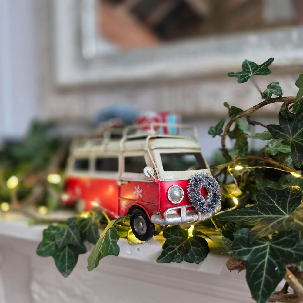 A small red and cream vintage-style camper van ornament decorated with a gray wreath and holiday boxes on top, displayed on a mantel surrounded by green ivy and warm string lights.