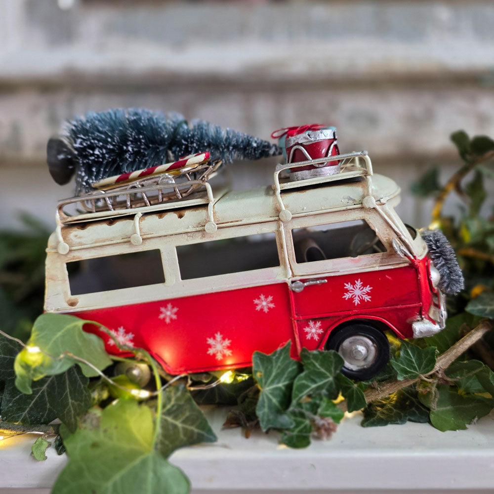 A decorative red and cream toy van decorated for Christmas, with snowflake patterns, a small artificial tree on the roof, and warm white string lights glowing from within, nestled among green ivy leaves.