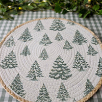 A round woven placemat with a quilted off-white surface featuring a repeating pattern of green pine trees, finished with a braided natural fiber rim, set on a green and white checkered tablecloth with blurred greenery in the background.