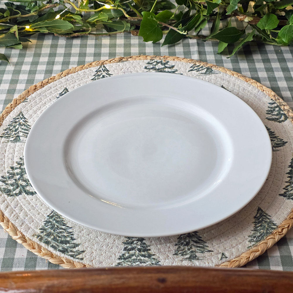 A white ceramic dinner plate layered on a round woven placemat with a green pine tree pattern, set on a green and white checkered tablecloth with green foliage garland in the background.