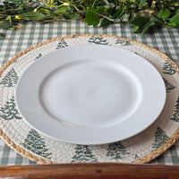 A white ceramic dinner plate layered on a round woven placemat with a green pine tree pattern, set on a green and white checkered tablecloth with green foliage garland in the background.