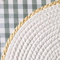 Close-up of a woven straw or raffia round mat with a braided natural fiber rim, placed against a green and white gingham checkered fabric background.