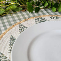 A close-up of a round white plate resting on a woven placemat with a green pine tree pattern, set on a green and white checkered tablecloth with green leaves at the top.