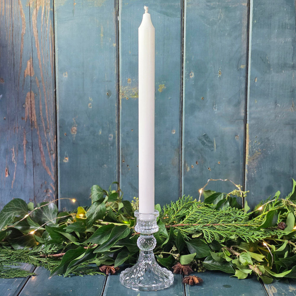 White candle in a glass holder on a wooden surface with greenery and string lights.
