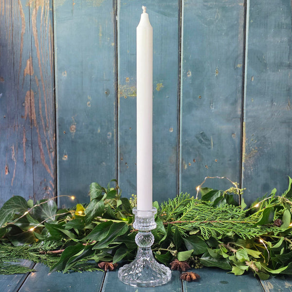 White candle in a glass holder on a wooden surface with greenery and string lights.
