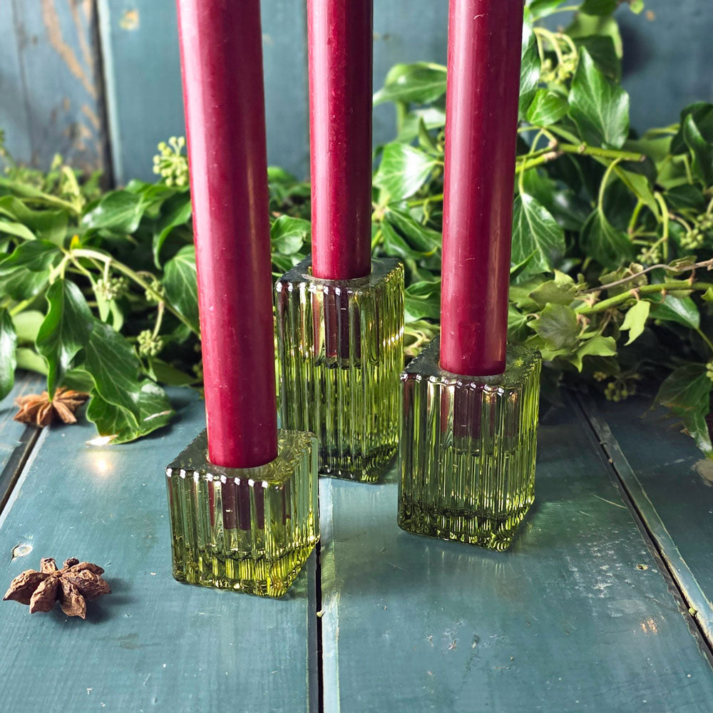 Three green candle holders with pink candles on a reflective surface with greenery in the background.
