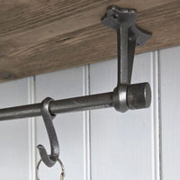 Highgate hanging hook and rod cap finial end, shown installed under a wooden shelf or cabinet against a white paneled wall.