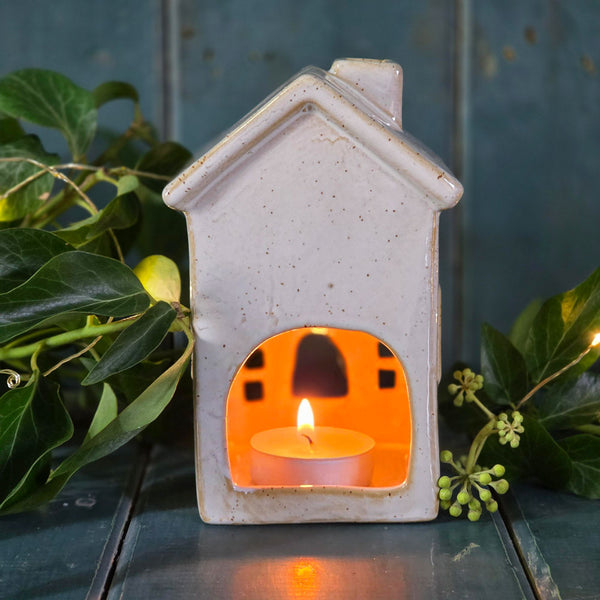 Ceramic house-shaped lantern with a lit candle inside, surrounded by green leaves on a wooden surface.