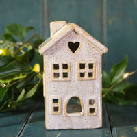 Ceramic house-shaped lantern with a heart window on a green surface with leaves and lights in the background.