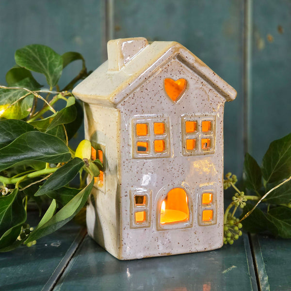 Ceramic house tea light holder lit by a tea light and surrounded by Christmas foliage.
