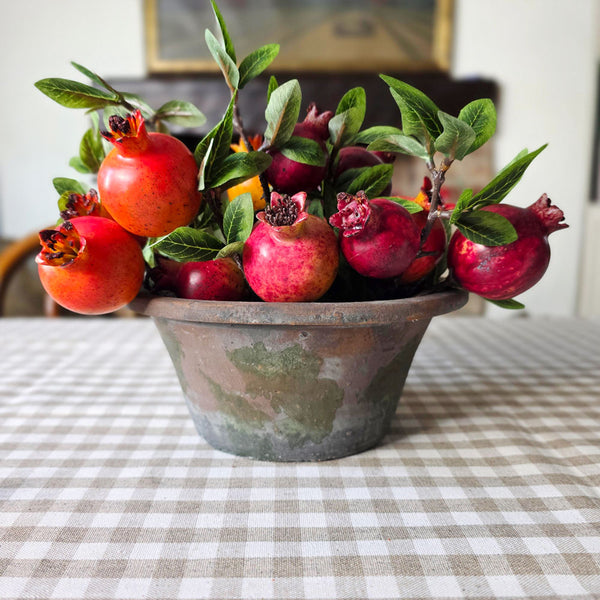 A decorative arrangement of red pomegranates with green leaves and stems, displayed in a rustic metal bowl on a checkered tablecloth in an indoor setting.