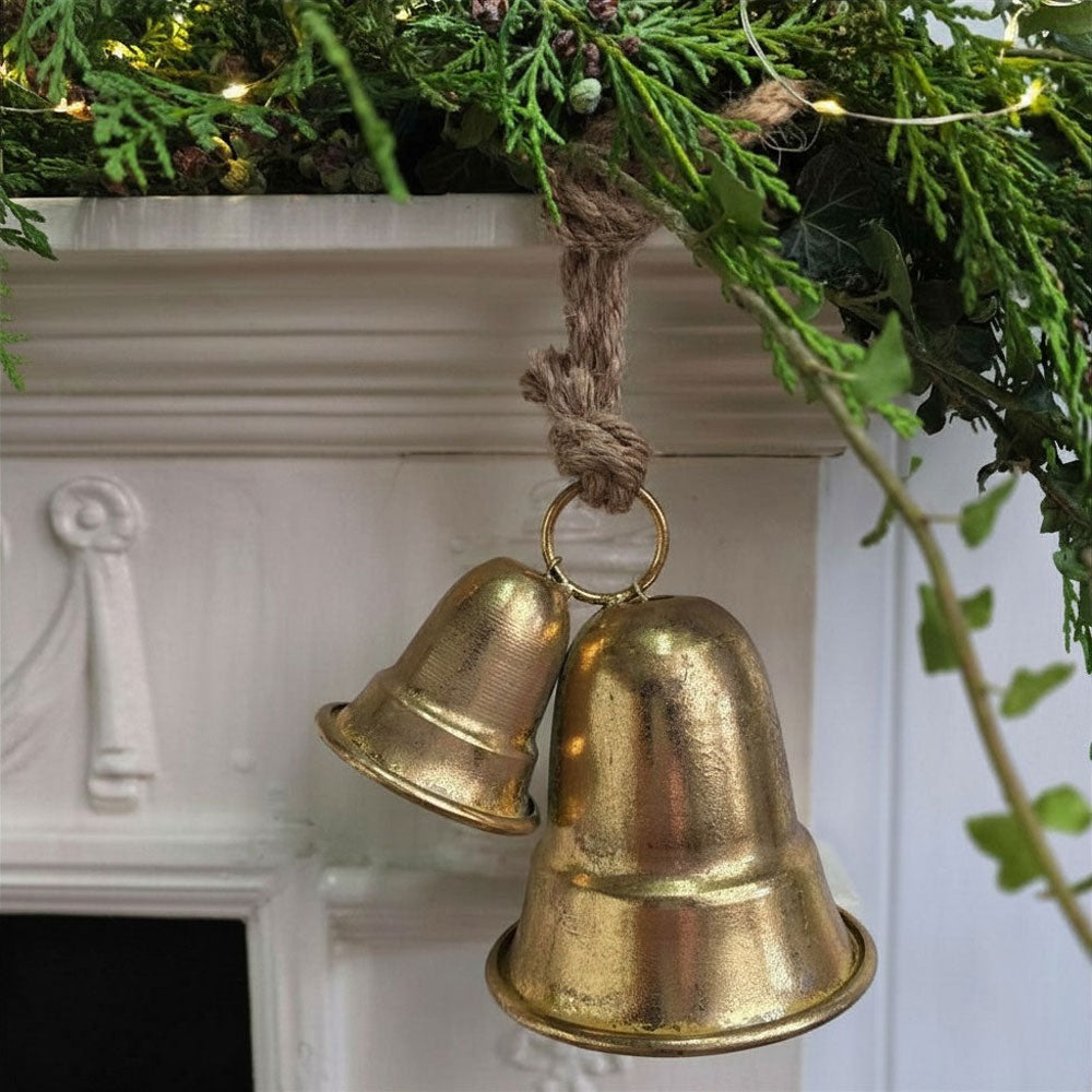 Two small metallic gold bells hanging from a rope ornament, displayed in front of a white fireplace mantel with green pine garland and warm string lights.