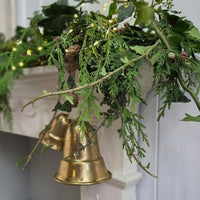 A decorative holiday garland with green evergreen branches, ivy leaves, small pinecones, and two hanging brass bells in front of a white fireplace mantel.