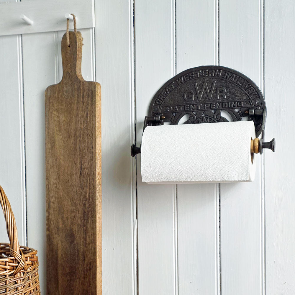 Wall-mounted paper towel holder with a wooden cutting board and wicker basket on a white paneled wall.