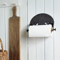 Wall-mounted paper towel holder with a wooden cutting board and wicker basket on a white paneled wall.