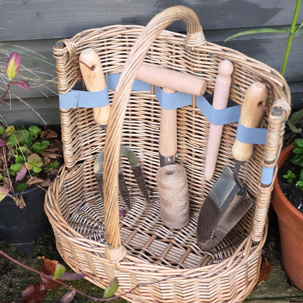 Wicker basket with gardening tools including shovels and a rake, surrounded by potted plants.