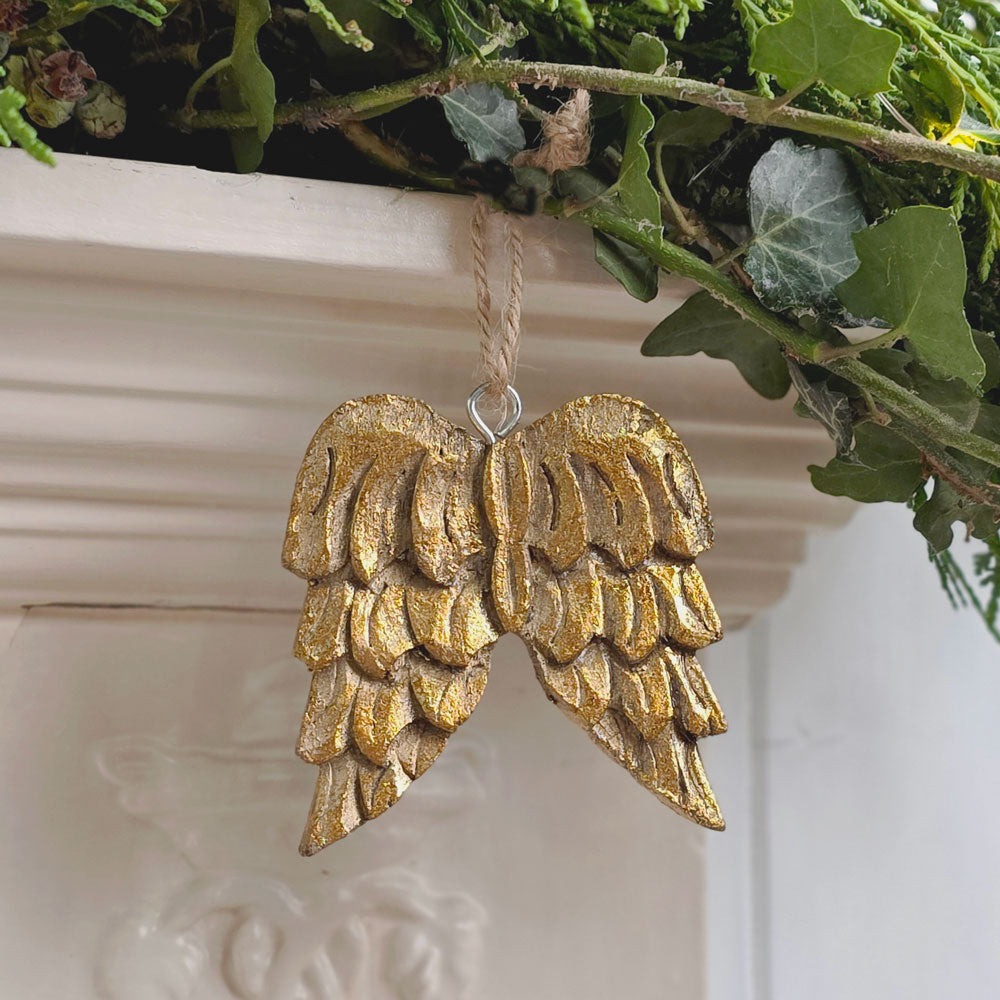 Wooden gold angel wings Christmas tree decoration hanging from a mantelpiece surrounded by foliage.
