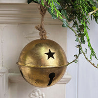 A vintage-style brass bell ornament with a star and keyhole cutout, hanging from a rope in front of a white fireplace mantel with green foliage.