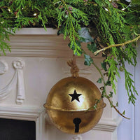 A gold decorative bell holiday ornament hanging from a white fireplace mantel decorated with green pine branches and ivy.
