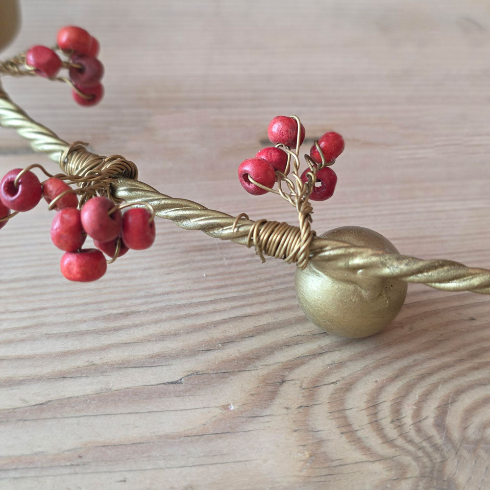 A close-up of a twisted gold metal branch decorated with small red berries and a round gold ball ornament, resting on a light wood surface.