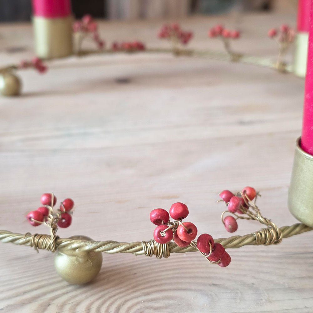 A close-up of a decorative wreath made from twisted gold wire and adorned with clusters of small red berries, shown on a light wood surface with candles partially visible in the background.