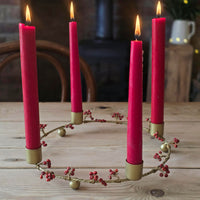 A circular Advent wreath with four lit pink taper candles in gold holders, displayed on a rustic wooden table.