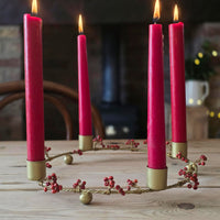 A set of four tall red taper candles in gold holders arranged in a circular wreath with red berries, lit and set on a wooden table in a cozy indoor setting.
