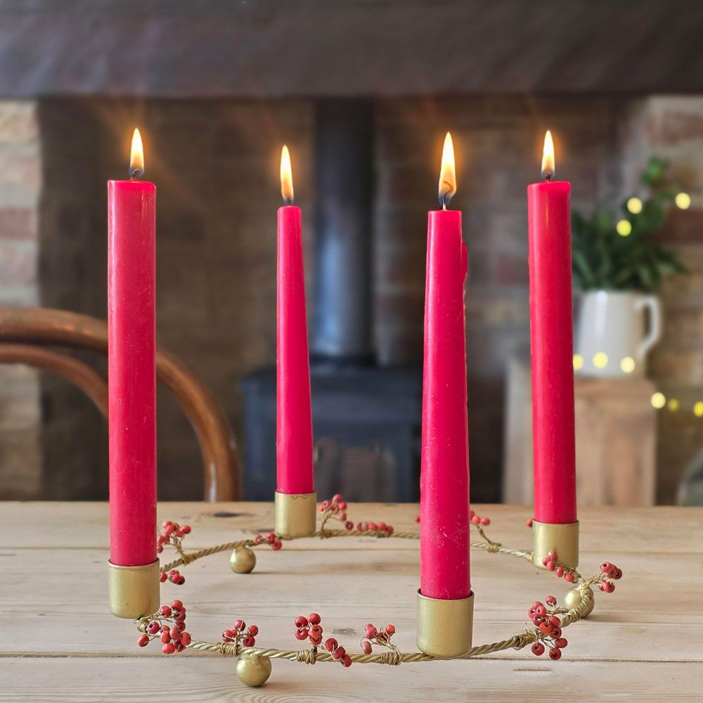 Four bright red taper candles in gold holders arranged in a circle on a wooden table, with a decorative gold and red berry garland around them. The candles are lit, and the background shows a cozy indoor setting with a fireplace and warm string lights.