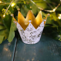 Unlit Decorative crown tea light holder with gold and silver accents on a green surface with foliage in the background.