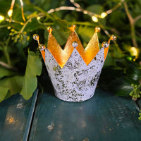 Decorative crown tea light holder with gold and white design on a green surface with leaves in the background